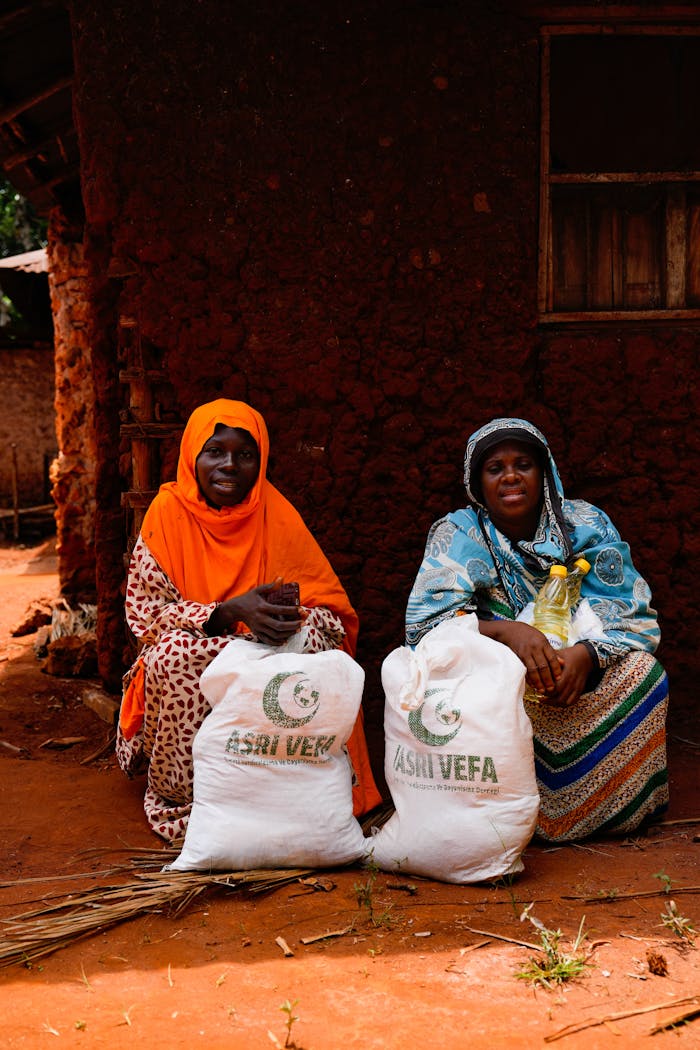 services-03 Two women sit by a mud house with relief bags, showing community aid.
